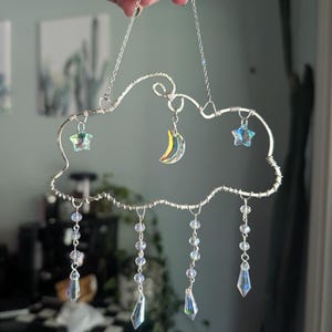 May include: A silver wire cloud-shaped suncatcher with hanging crystal beads and charms. The suncatcher features a crescent moon and two stars, all with iridescent finishes. The cloud has a chain for hanging.