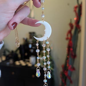 May include: A gold-coloured necklace with a white crescent moon pendant. The moon has a profile design and is suspended from a gold chain. Clear beads and iridescent teardrop crystals dangle from the moon. The necklace is held up by a hand.