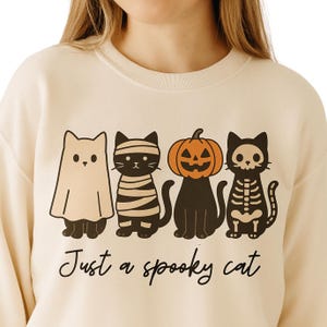 May include: Cream-colored sweatshirt featuring a Halloween-themed graphic of four cartoon cats dressed as a ghost, mummy, jack-o'-lantern, and skeleton. The phrase "Just a spooky cat" is written below the cats in black script.