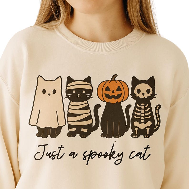 May include: Cream-colored sweatshirt featuring a Halloween-themed graphic of four cartoon cats dressed as a ghost, mummy, jack-o'-lantern, and skeleton. The phrase "Just a spooky cat" is written below the cats in black script.
