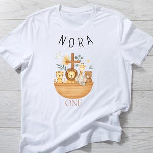 May include: White t-shirt featuring a watercolor illustration of an ark with animals, a cross, and the name "NORA" above. The word "ONE" is below the ark. The design includes a lion, giraffe, tiger, elephant, and bear.