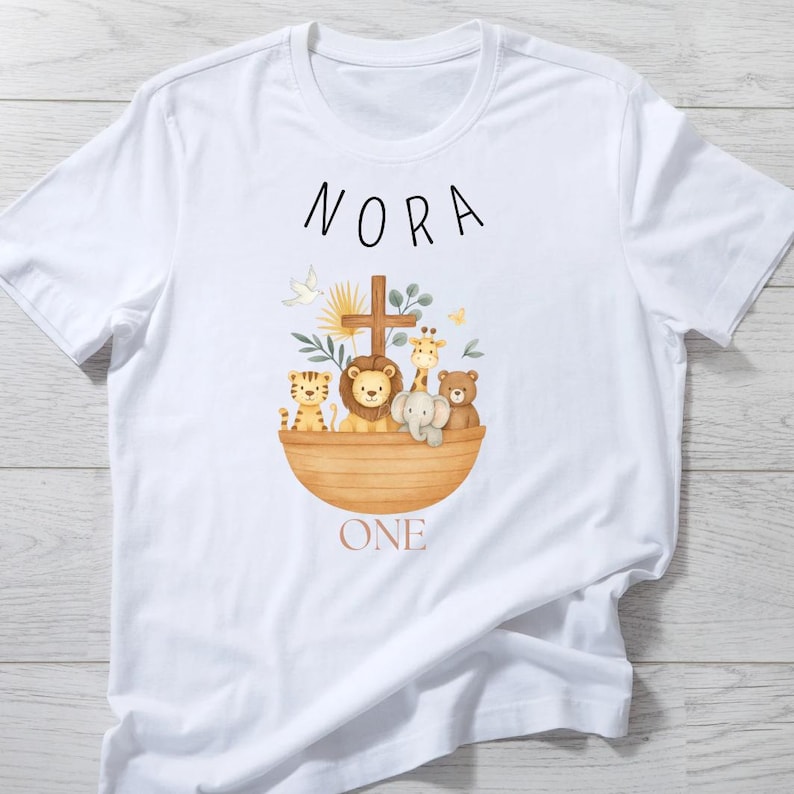 May include: White t-shirt featuring a watercolor illustration of an ark with animals, a cross, and the name "NORA" above. The word "ONE" is below the ark. The design includes a lion, giraffe, tiger, elephant, and bear.