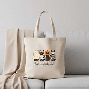 May include: Beige tote bag featuring a Halloween-themed design. The graphic includes four cartoon cats dressed as a ghost, mummy, pumpkin, and skeleton. The text "Just a spooky cat" is printed below the cats.