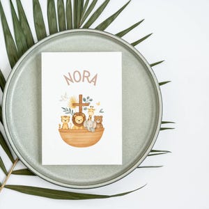 May include: A white card with the name "NORA" in light brown lettering. The card features an illustration of Noah's Ark with various animals, including a tiger, lion, giraffe, elephant, and bear. The card is on a gray plate with green leaves.