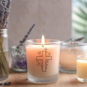 Op de afbeelding: Een brandende, perzik kleurige kaars in een heldere glazen pot. Een zwart kruisontwerp met bloemaccenten is op de voorkant gedrukt. Andere kaarsen en lavendel takjes in glazen potten staan op de achtergrond.