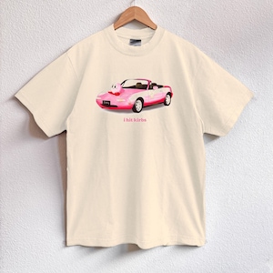 May include: Cream-colored t-shirt featuring a pink convertible car graphic with a cartoon character. The car has white accents and red wheels. The text "i hit kirbs" is printed below the car. The shirt is hanging on a wooden hanger.