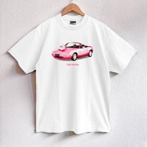 May include: White t-shirt featuring a pink convertible car graphic with a cartoon character and the text "i hit kirbs". The car has white accents and red wheels. The shirt is hanging on a wooden hanger.