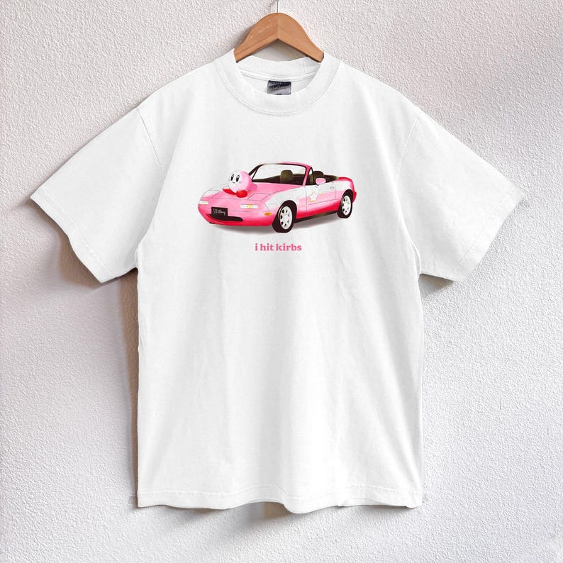 May include: White t-shirt featuring a pink convertible car graphic with a cartoon character and the text "i hit kirbs". The car has white accents and red wheels. The shirt is hanging on a wooden hanger.