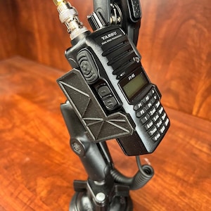 Puede incluir: Una radio transceptor de banda dual Yaesu FT-65 negra montada sobre una base de ventosa negra. La radio tiene una pequeña pantalla y un teclado. Una antena está conectada a la parte superior de la radio.