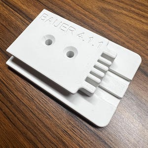 May include: A white, 3D-printed object with the text "BAUER 4.1.1" on the top surface. The object has two circular holes and a series of rectangular cutouts. It is resting on a brown wooden surface.