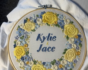 May include: Embroidered hoop art with a wreath of yellow and blue flowers surrounding the names "Kylie & Jace" in blue cursive lettering.