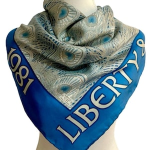 May include: A square silk scarf with a peacock feather design in shades of blue and silver. The scarf has a bright blue border with the word "LIBERTY" in white and the year "1981".
