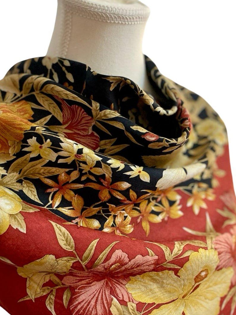 May include: A patterned scarf with a floral design. The scarf has a black background with yellow and orange flowers, and a red border with large yellow hibiscus flowers. The scarf is draped over a mannequin.