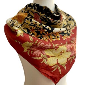 May include: A silk scarf with a vibrant floral design. The scarf features a red border with large yellow and pink hibiscus flowers. The centre has a black background with smaller yellow and orange flowers. The scarf is draped over a mannequin.