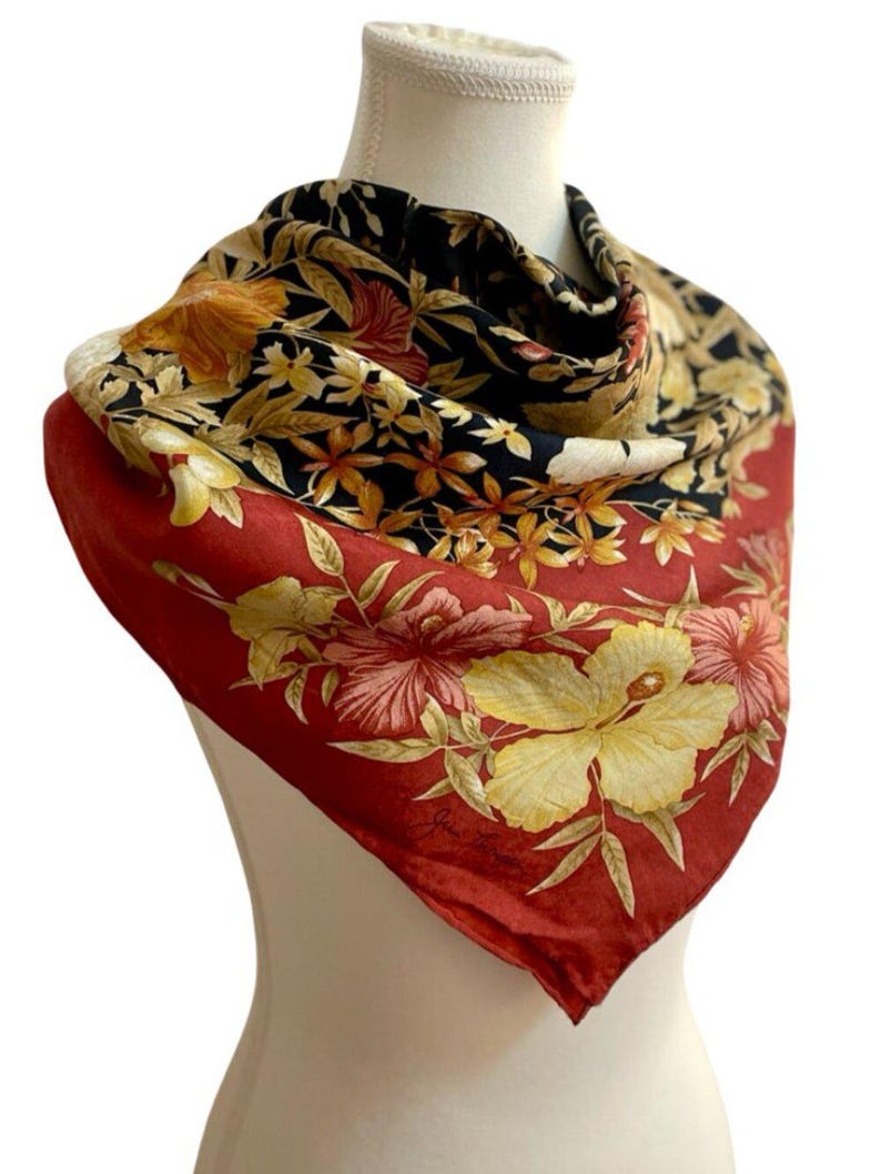 May include: A silk scarf with a vibrant floral design. The scarf features a red border with large yellow and pink hibiscus flowers. The centre has a black background with smaller yellow and orange flowers. The scarf is draped over a mannequin.