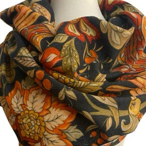 May include: A scarf featuring a dark charcoal background with a repeating pattern of large orange and yellow flowers, green leaves, and small berries. The fabric is draped around a mannequin neck, displaying the intricate botanical print throughout.