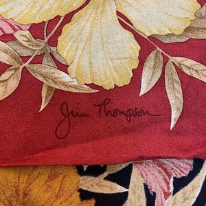 May include: Silk scarf with a floral pattern. The design includes large yellow flowers, pink details, and brown leaves on a red backdrop. The signature "Jim Thompson" is present. The lower section of the scarf has a black background with yellow and orange flowers.