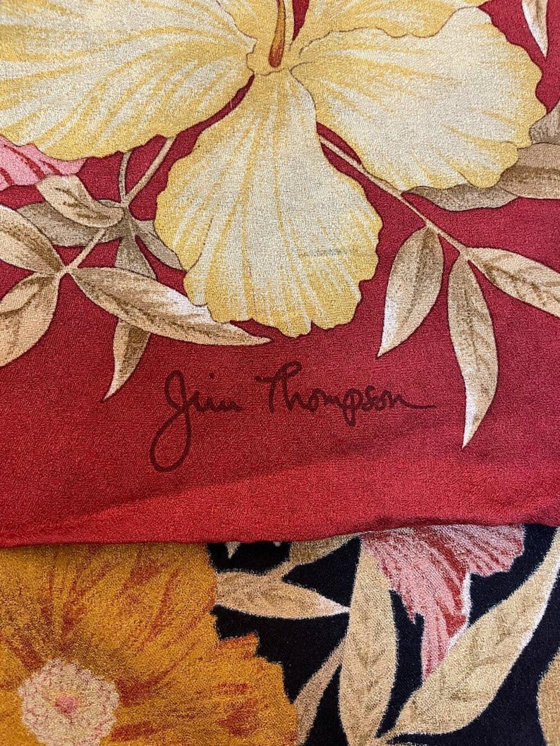 May include: Silk scarf with a floral pattern. The design includes large yellow flowers, pink details, and brown leaves on a red backdrop. The signature "Jim Thompson" is present. The lower section of the scarf has a black background with yellow and orange flowers.