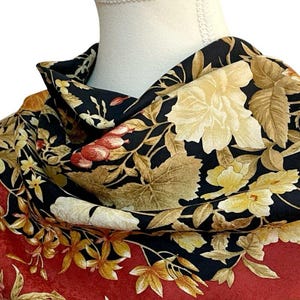 May include: A floral scarf with a black background and a red border. The scarf features large, cream-coloured flowers and smaller yellow and red blossoms. The design includes detailed leaves in shades of brown and gold.