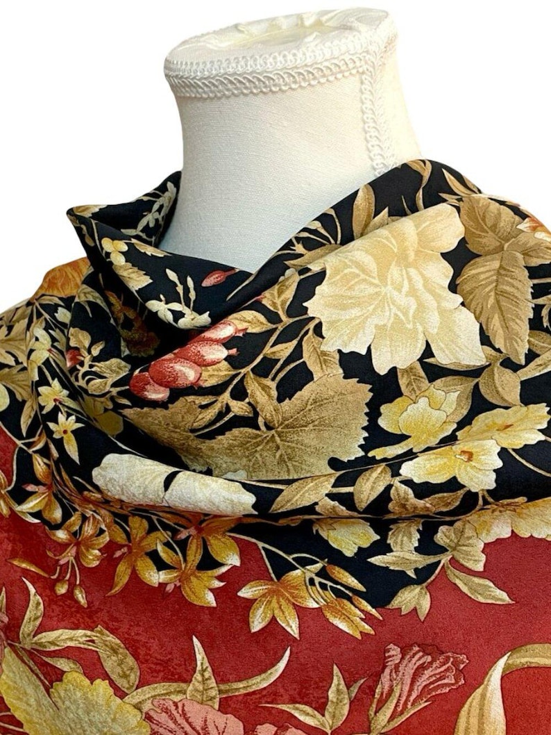 May include: A floral scarf with a black background and a red border. The scarf features large, cream-coloured flowers and smaller yellow and red blossoms. The design includes detailed leaves in shades of brown and gold.