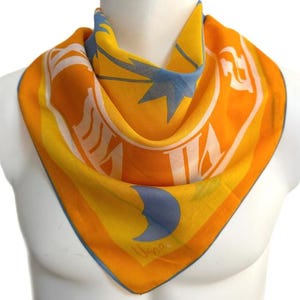 May include: A square scarf in shades of orange, yellow, and blue, featuring a star and crescent moon design. The scarf is draped around a mannequin's neck, showcasing its vibrant colours and intricate pattern. The scarf has a blue border.