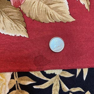 May include: A close-up of a fabric with a floral pattern. The top portion features large red roses and tan leaves on a red background. The bottom portion has a black background with gold-coloured leaves. A silver coin is on the red fabric.