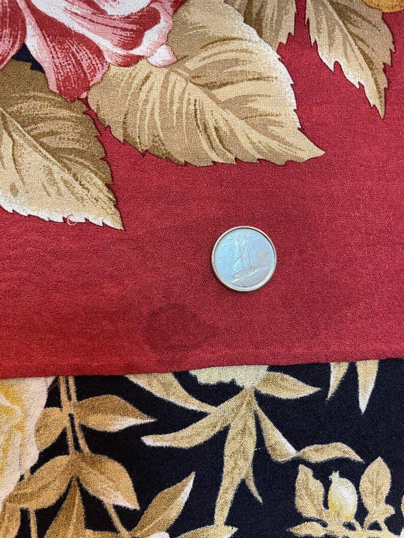 May include: A close-up of a fabric with a floral pattern. The top portion features large red roses and tan leaves on a red background. The bottom portion has a black background with gold-coloured leaves. A silver coin is on the red fabric.