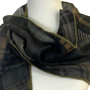 May include: A scarf featuring a dark plaid pattern in shades of charcoal grey, black, and olive green. The fabric has a soft, textured appearance with a distinct olive green stitched border along the edges, draped around a white mannequin neck.