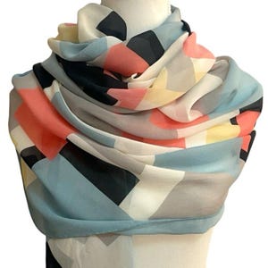 May include: A lightweight scarf featuring a geometric pattern of overlapping shapes in shades of coral, dusty blue, black, cream, and pale yellow. The fabric is draped in multiple layers around a mannequin neck, displaying a soft, semi-sheer texture.
