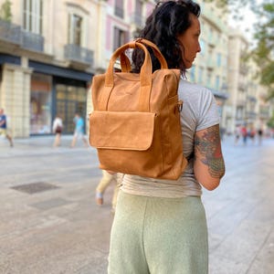 Peut inclure: Sac à dos en cuir de couleur beige avec une poche avant et des poignées supérieures. Le sac est porté sur l'épaule. L'arrière-plan montre une rue de la ville avec des bâtiments et des personnes.