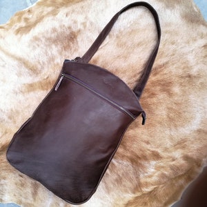 Peut inclure: Un sac à bandoulière en cuir marron foncé doté d'une longue sangle unique et d'une poche zippée horizontale sur la partie supérieure avant. Le sac présente un bord supérieur incurvé et un corps rectangulaire aux coins inférieurs arrondis.