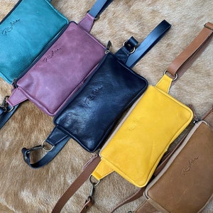Puede incluir: Cinco riñoneras de cuero en diferentes colores: verde azulado, burdeos, negro, amarillo y marrón. Cada riñonera tiene una cremallera y una pequeña etiqueta de cuero con la palabra "Rishia" grabada.