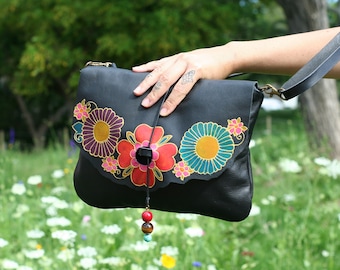 May include: Black leather shoulder bag with a colorful floral design. The flowers are painted in shades of red, purple, yellow, and teal. The bag has a black strap and a decorative tassel with beads.