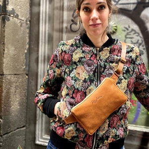 May include: A brown leather bum bag with a gold buckle and a strap. The bum bag is worn by a person wearing a floral patterned jacket.