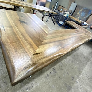 Può includere: Un grande tavolo in legno fatto a mano con un bordo naturale e un design angolare a forma di V. Il tavolo è realizzato in legno marrone chiaro con venature visibili e una finitura liscia e lucida. Le dimensioni del tavolo non sono visibili.