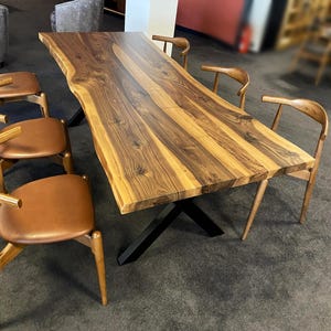 Könnte beinhalten: Ein großer, rechteckiger Esstisch aus Holz mit einer Baumkante und einem dunklen, kreuzförmigen Metallfuß. Der Tisch ist von mehreren Holzstühlen mit braunen Ledersitzen umgeben. Die Holzmaserung ist deutlich sichtbar.