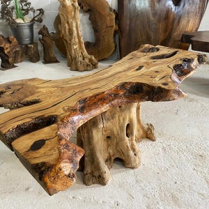 May include: A rustic wooden coffee table with a live edge top and a natural wood finish. The table has a unique, irregular shape and is supported by two large, natural wood legs.