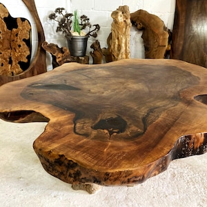 Puede incluir: Una mesa de centro de madera grande, de forma irregular, con borde natural. La mesa está hecha de una sola losa de madera, mostrando la veta y las variaciones de color. La mesa mide aproximadamente 152 cm de largo y 102 cm de ancho.