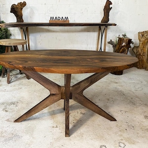 May include: A large, round wooden table with a dark brown finish. The table has a unique starburst-style base made of intersecting wooden beams. The table is a centerpiece in a room with other wooden furniture and decorative elements.