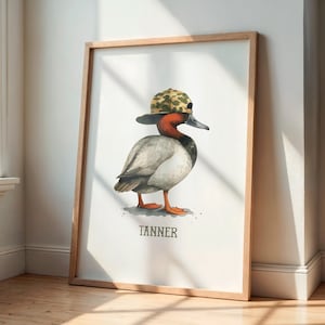 May include: Watercolor print of a duck wearing a camouflage baseball cap. The duck has a red head and a gray body. The name "TANNER" is printed in black below the duck. The artwork is framed in a light brown wooden frame.