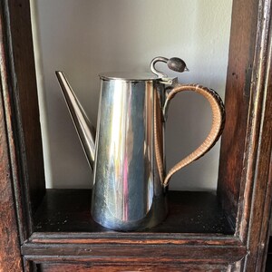 Peut inclure: Une cafetière en argent poli avec un long bec et un couvercle. La poignée est enveloppée d'un matériau brun clair. Le pot a un élément décoratif sur le couvercle. Le pot est exposé sur une étagère en bois foncé.