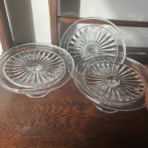 May include: Three clear glass serving dishes with a raised base and a sunburst design. The dishes are arranged on a dark wooden surface, with a wooden cabinet in the background. The glass dishes are likely vintage.