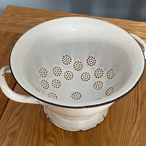 Op de afbeelding: Een witte emaille vergiet met een zwarte rand en twee handvatten. De vergiet heeft talrijke kleine gaatjes om af te wateren en staat op een voetstuk. Een klassiek keukengereedschap om voedsel te wassen en af te gieten.