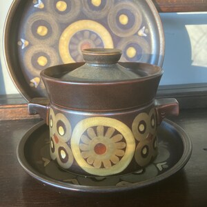 May include: A vintage brown ceramic serving dish set with a lid, handles, and a matching plate. The set features a repeating pattern of yellow and brown circles and floral designs. The set is likely from the mid-century era.