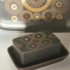 May include: A rectangular, dark grey butter dish with a matching lid. The lid and dish are decorated with a repeating pattern of brown and yellow circles with red accents. The dish sits on a brown base.