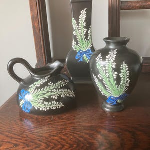 May include: Three black ceramic vases with floral designs. The vases are decorated with white and green floral patterns and blue bows. The vases are arranged on a wooden surface.