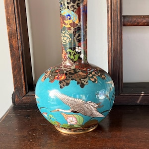 May include: A cloisonné vase with a turquoise blue base featuring a white crane in flight, pink flowers, and green leaves. The neck of the vase is decorated with a multi-coloured geometric and floral pattern in shades of black, red, and gold.