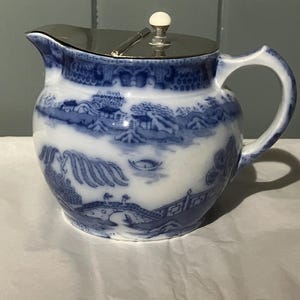 Può includere: Una lattiera in ceramica blu e bianca con coperchio argentato. La lattiera presenta un design panoramico con ponti, alberi ed edifici. Il manico è curvo e il coperchio ha un piccolo pomello bianco.