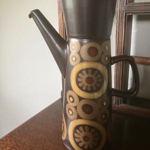 May include: A brown ceramic coffee pot with a tall, cylindrical body and a long spout. The pot features a repeating pattern of yellow and brown circles and flower-like shapes. It has a handle and a lid.