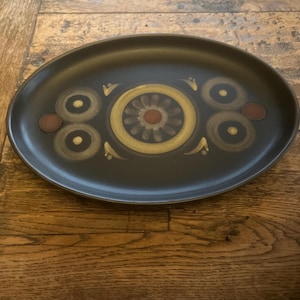 May include: A dark brown oval ceramic platter featuring a central design of gold, brown, and red concentric circles. The platter has a matte finish and is placed on a wooden surface. The design is symmetrical and geometric.